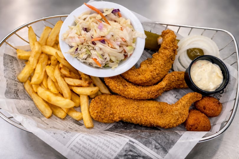 Fried fish and hush puppies with fries, coleslaw, and tartar sauce in a metal basket.