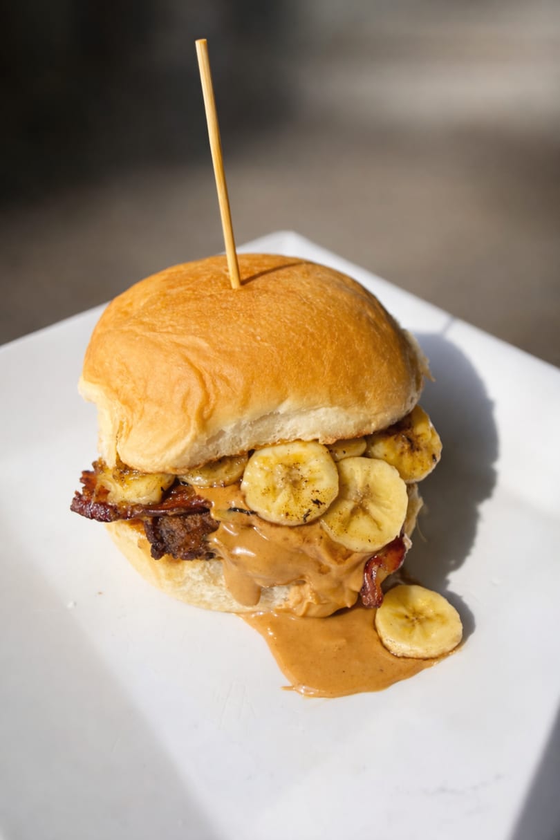 A peanut butter, bacon, and banana sandwich on a bun, with peanut butter dripping onto a white plate.